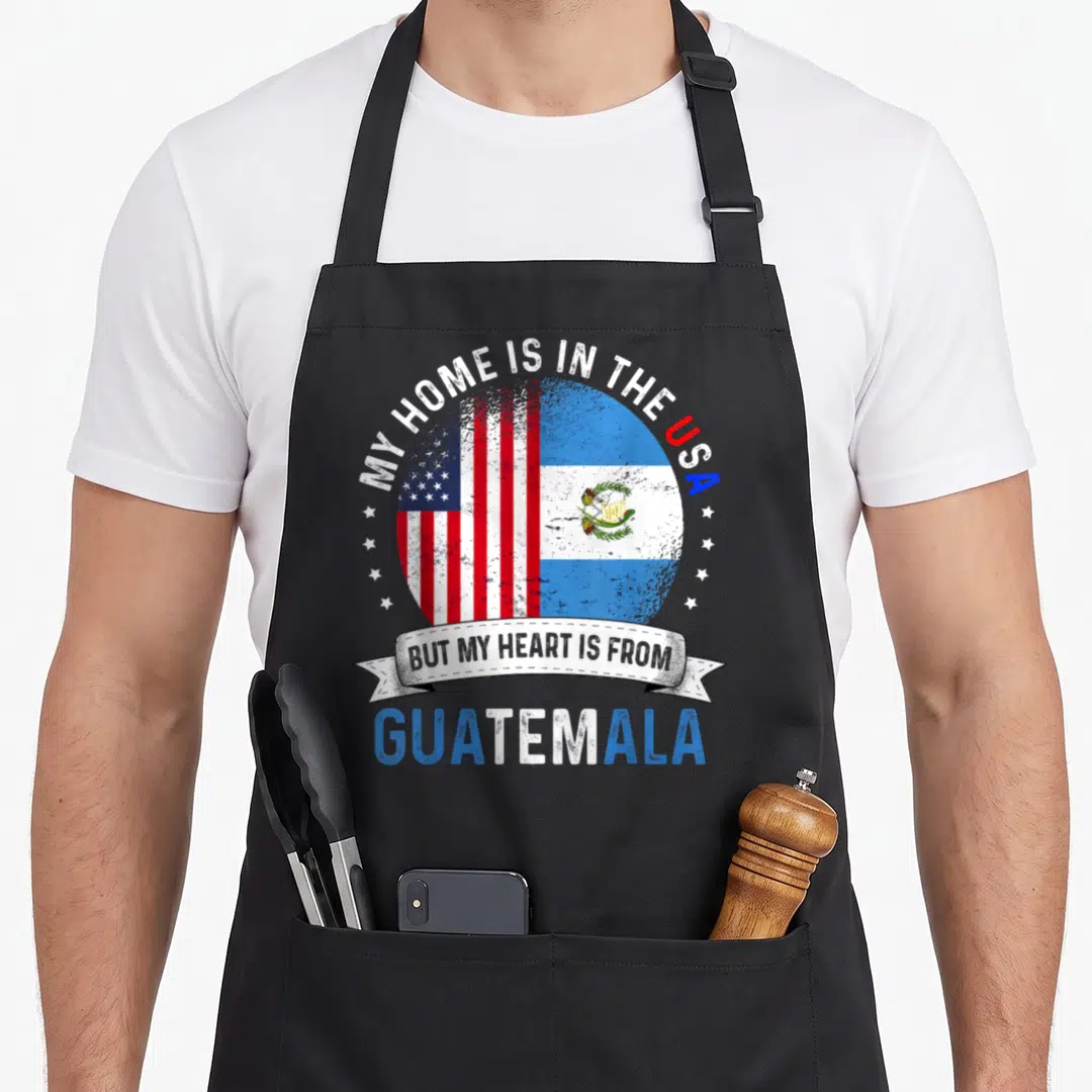 Guatemalan american flag apron - Split circle US and Guatemala flags with arched text MY HOME IS IN THE USA — shown on a black cotton canvas apron against a white background