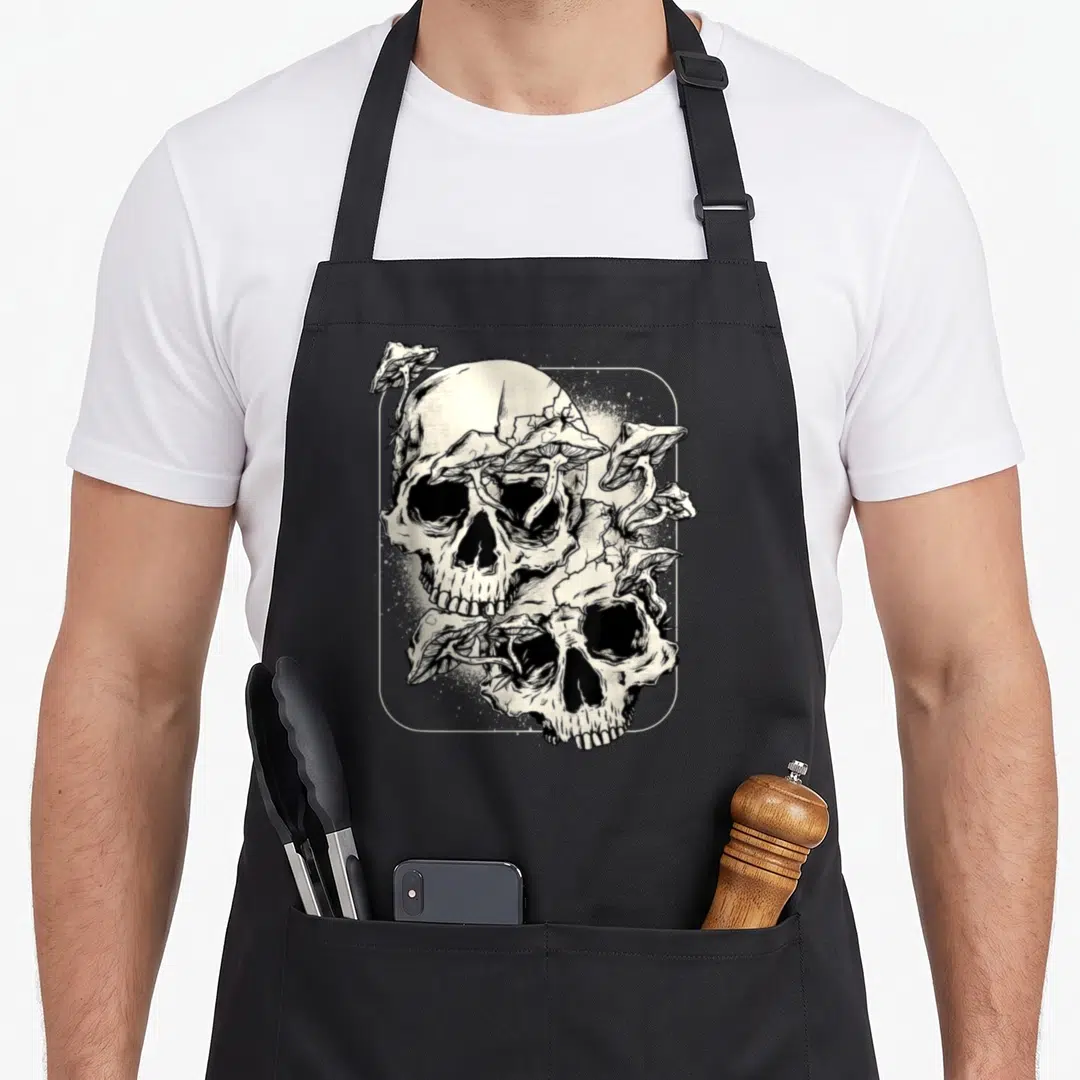 Skull goth apron - Two skulls stacked vertically with mushrooms growing from them inside a thin white frame — shown on a black cotton canvas apron against a white background