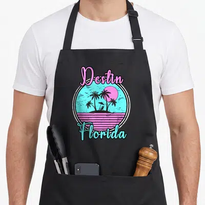 Travel florida apron - Retro circle with Destin Florida text, palm trees, sun, and beach chair in pink and blue — shown on a black cotton canvas apron against a white background