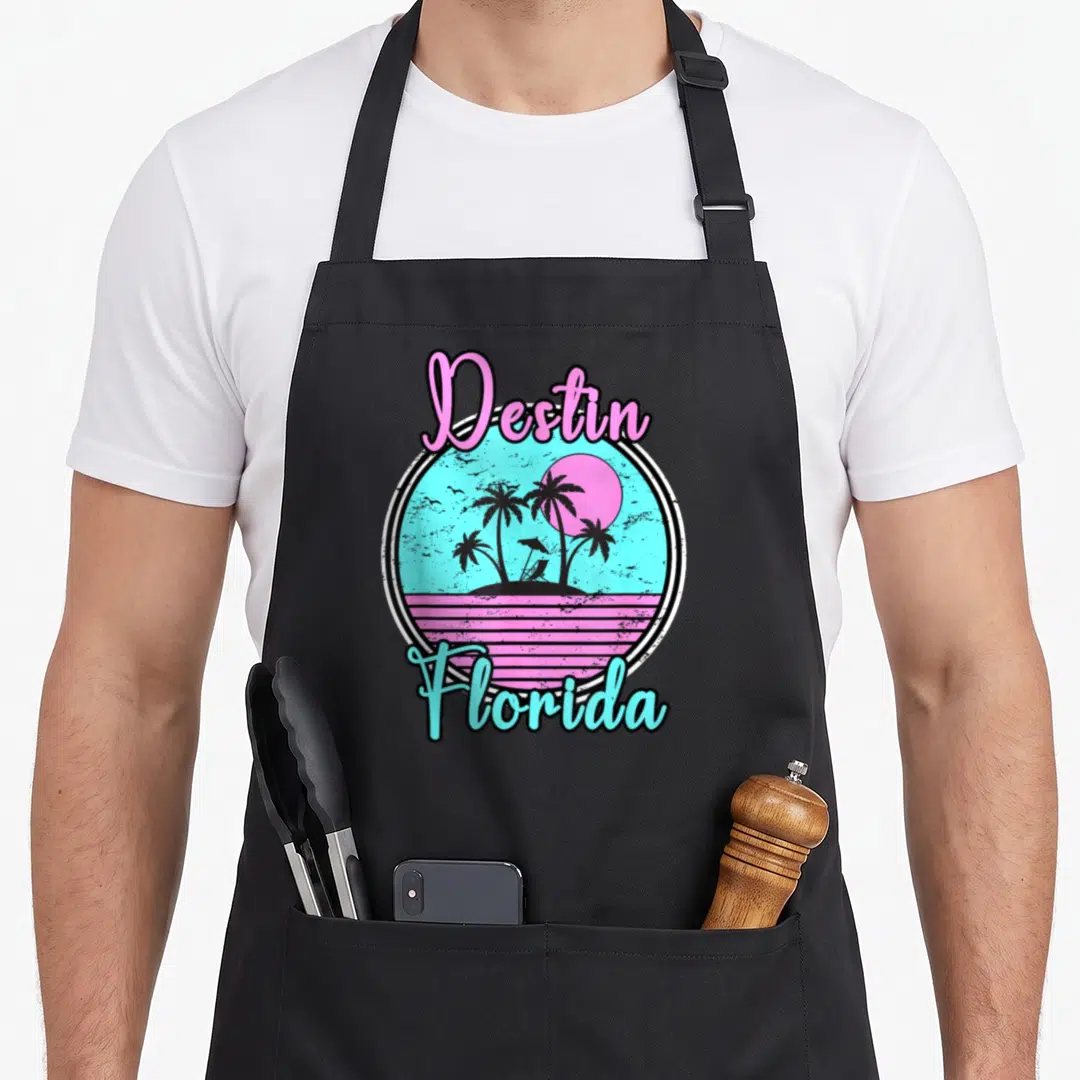 Travel florida apron - Retro circle with Destin Florida text, palm trees, sun, and beach chair in pink and blue — shown on a black cotton canvas apron against a white background