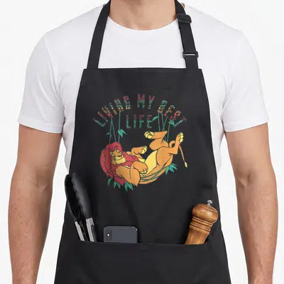 Living lion apron - Cartoon lion lying on back under arched text LIVING MY BEST LIFE with teal leaves — shown on a black cotton canvas apron against a white background