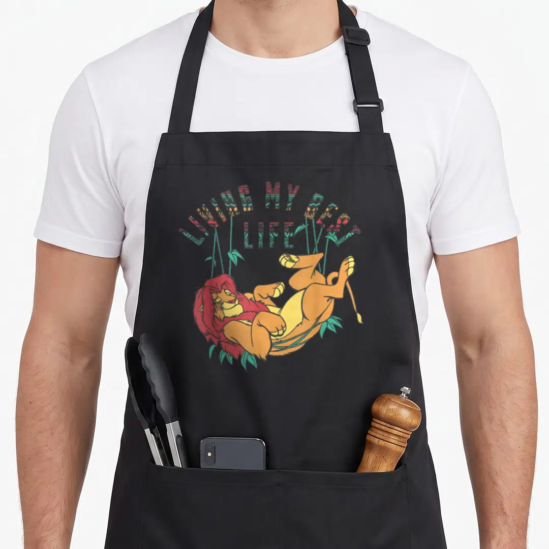 Living lion apron - Cartoon lion lying on back under arched text LIVING MY BEST LIFE with teal leaves — shown on a black cotton canvas apron against a white background