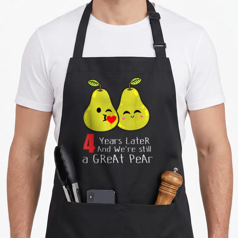 4th wedding aprons - Two cartoon yellow pears kissing a red heart above 4 Years Later Great Pear text — shown on a black cotton canvas apron against a white background