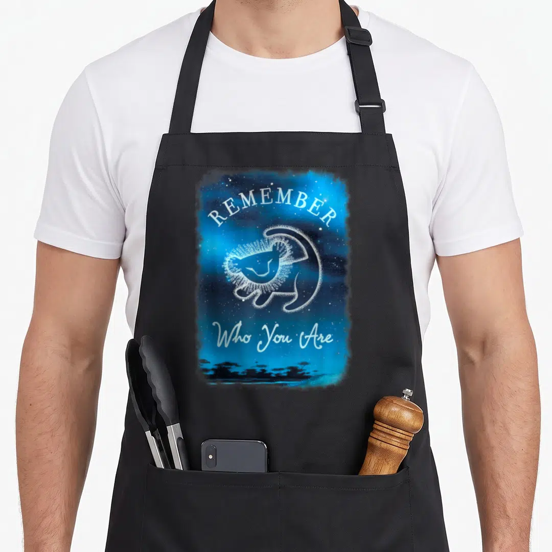 Remember constellation apron - White starry lion silhouette on blue background with REMEMBER Who You Are text and tree — shown on a black cotton canvas apron against a white background