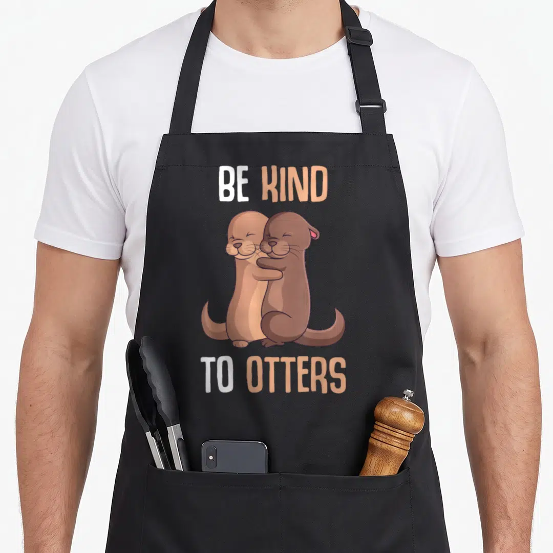 Kind otter mom apron - Two cartoon otters hugging with BE KIND TO OTTERS text in brown white peach colors — shown on a black cotton canvas apron against a white background