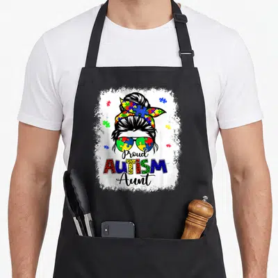 Autism messy aprons - Cartoon woman bun puzzle sunglasses Proud Autism Aunt text — shown on a black cotton canvas apron against a white background