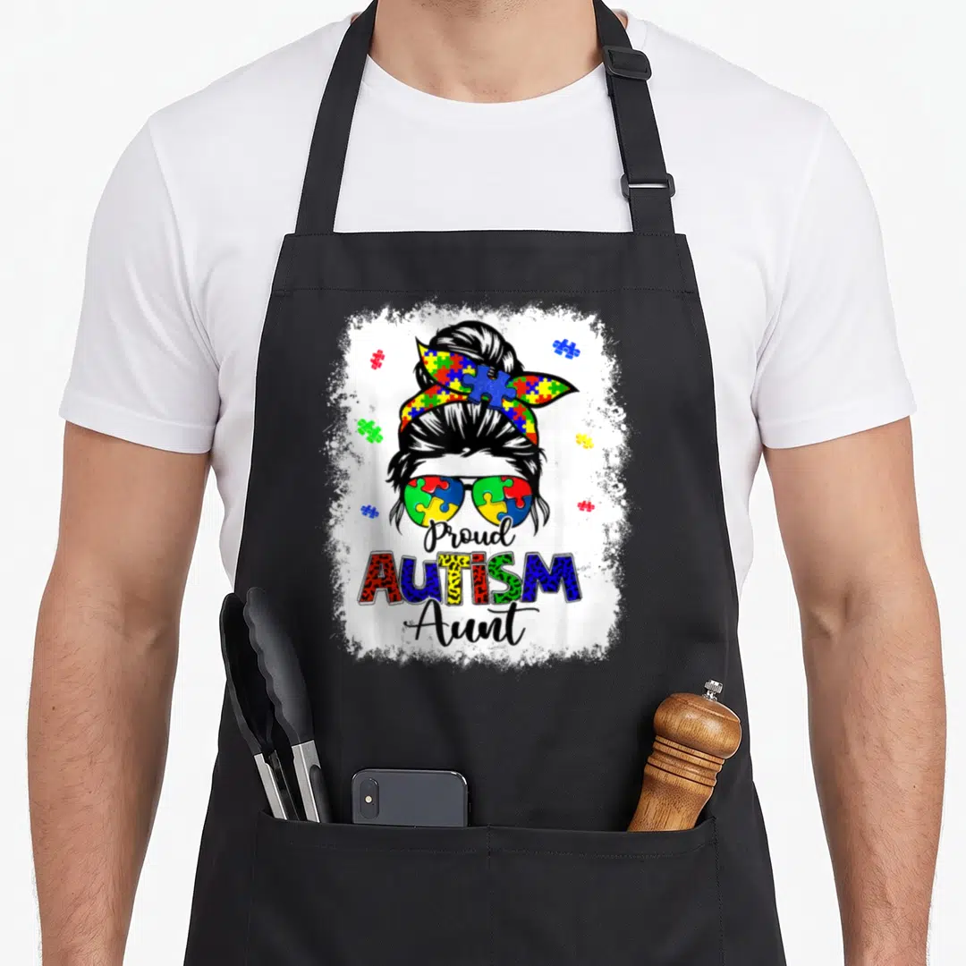 Autism messy aprons - Cartoon woman bun puzzle sunglasses Proud Autism Aunt text — shown on a black cotton canvas apron against a white background
