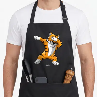 Dabbing tiger apron - Cartoon tiger in orange black white doing a dab pose on black apron — shown on a black cotton canvas apron against a white background