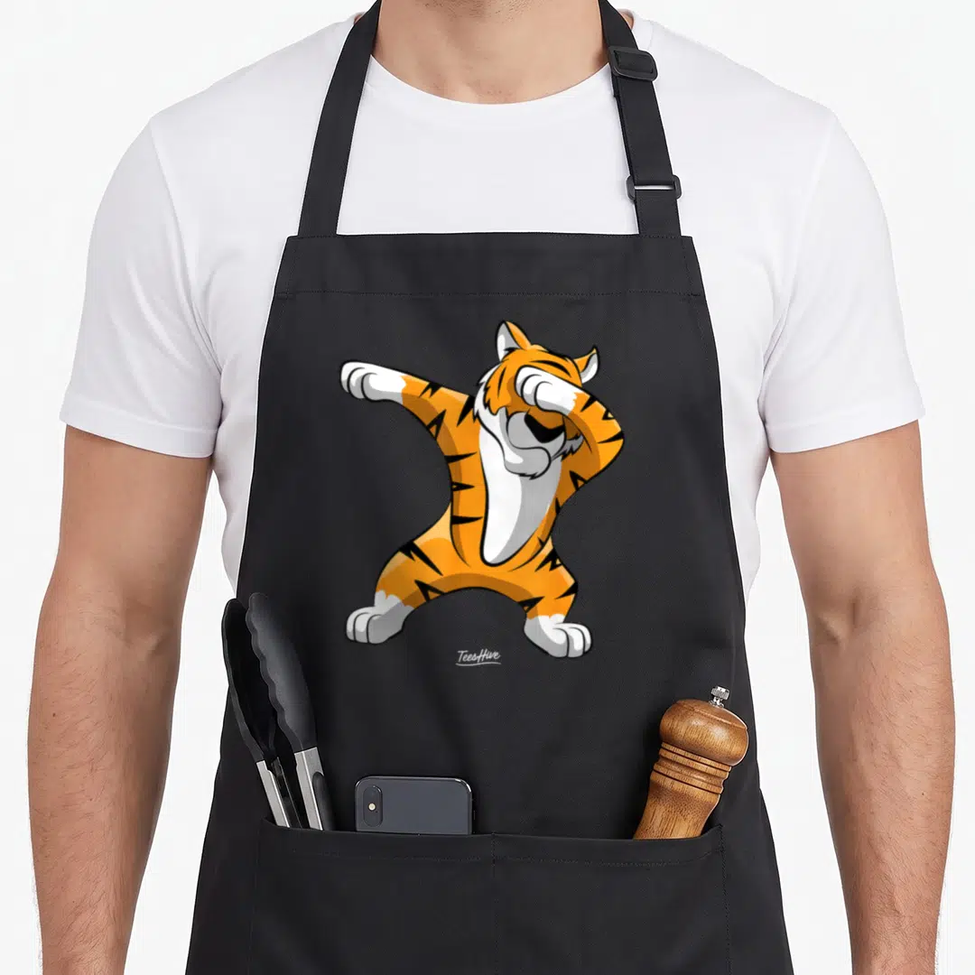 Dabbing tiger apron - Cartoon tiger in orange black white doing a dab pose on black apron — shown on a black cotton canvas apron against a white background