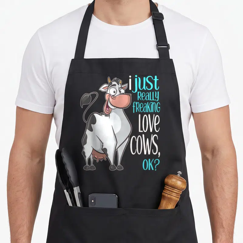 Freaking cow apron - Cartoon cow standing next to stacked text reading i just really freaking love cows ok in cyan — shown on a black cotton canvas apron against a white background