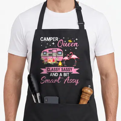 Queen smart apron - Pink camper trailer with two flamingos, campfire, flowers, and smart assy text — shown on a black cotton canvas apron against a white background