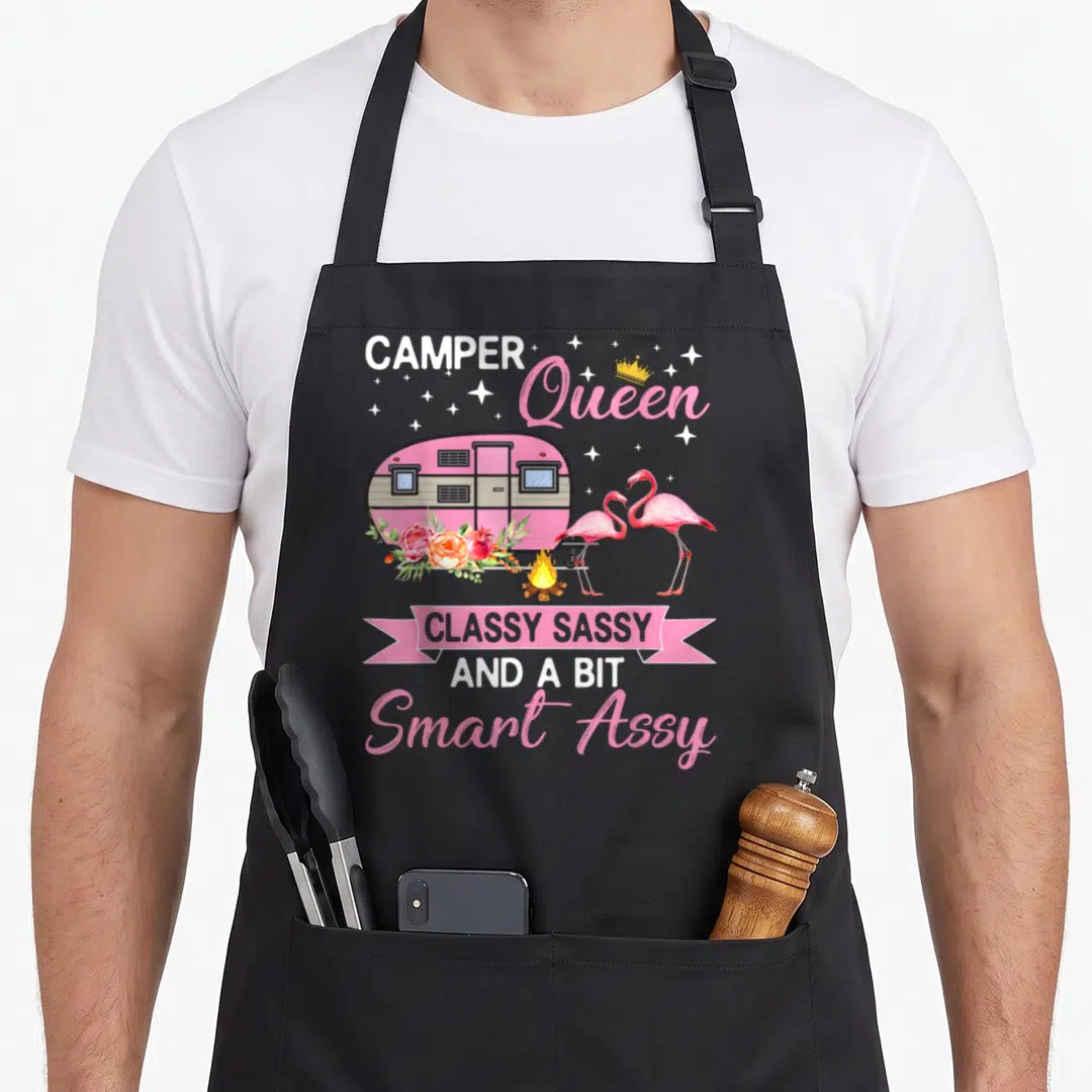 Queen smart apron - Pink camper trailer with two flamingos, campfire, flowers, and smart assy text — shown on a black cotton canvas apron against a white background