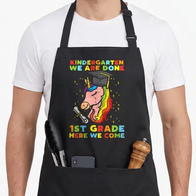 Grade unicorn aprons - Cartoon unicorn with graduation cap diploma and rainbow text reading kindergarten we are done — shown on a black cotton canvas apron against a white background