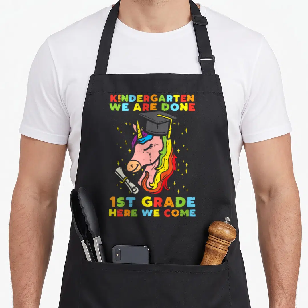Grade unicorn aprons - Cartoon unicorn with graduation cap diploma and rainbow text reading kindergarten we are done — shown on a black cotton canvas apron against a white background