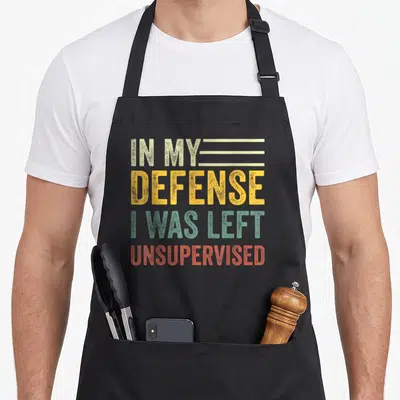 Funny sarcastic left apron - Stacked text IN MY DEFENSE I WAS LEFT UNSUPERVISED in beige yellow green and red — shown on a black cotton canvas apron against a white background