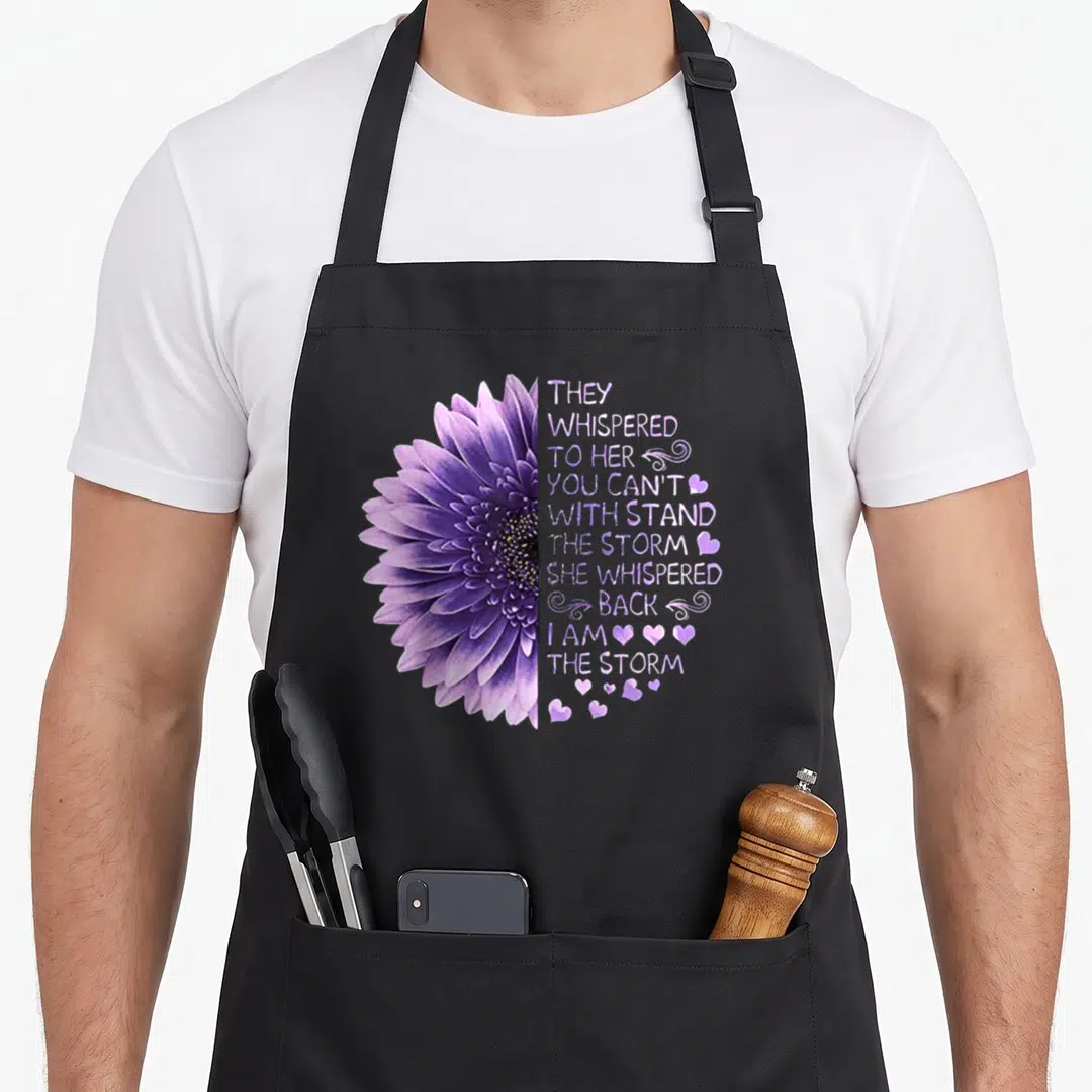 They her apron - Realistic purple flower next to stacked text They Whispered To Her You Can't Withstand The Storm — shown on a black cotton canvas apron against a white background