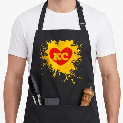 Funny kansas girl apron - Distressed KC text inside a red heart on a yellow paint splatter — shown on a black cotton canvas apron against a white background