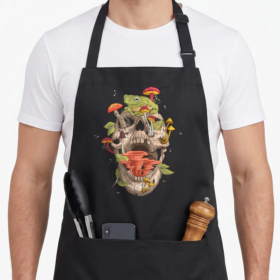 Skull cottagecore apron - Beige skull with green frogs, red and orange mushrooms, and leaves sprouting from eye sockets — shown on a black cotton canvas apron against a white background