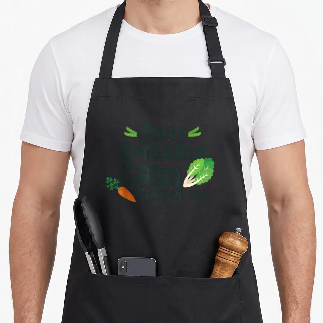 Peas romaine calm and carrot on apron - Stacked text Peas Romaine Calm Carrot with illustrated pea pods lettuce leaf — shown on a black cotton canvas apron against a white background