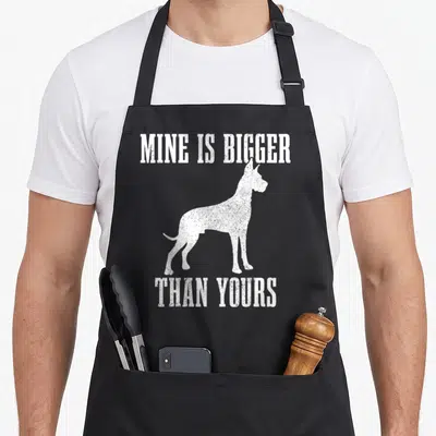 Mine dog apron - White distressed Great Dane silhouette between text MINE IS BIGGER THAN YOURS — shown on a black cotton canvas apron against a white background