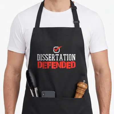 Dissertation boredkoalas apron - White checkbox with red check mark above distressed DISSERTATION DEFENDED text — shown on a black cotton canvas apron against a white background