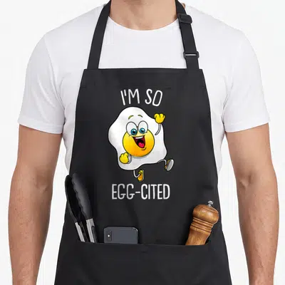 Pun egg apron - Running cartoon fried egg with I'M SO EGG-CITED text in white yellow blue and red — shown on a black cotton canvas apron against a white background