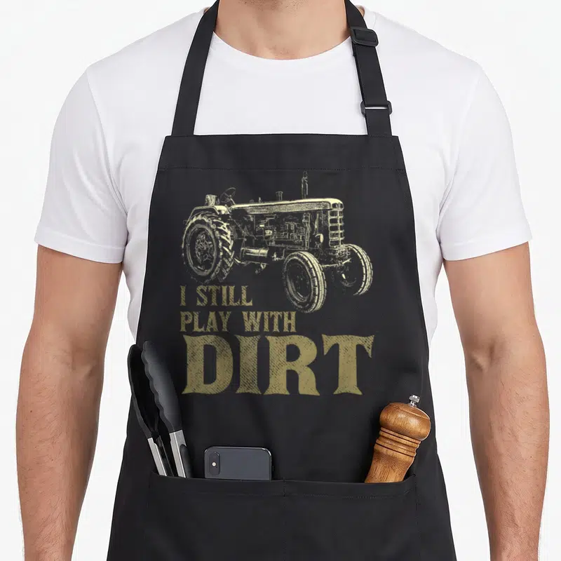 Play farm apron - Beige tractor illustration above three lines of distressed text reading I STILL PLAY WITH DIRT — shown on a black cotton canvas apron against a white background