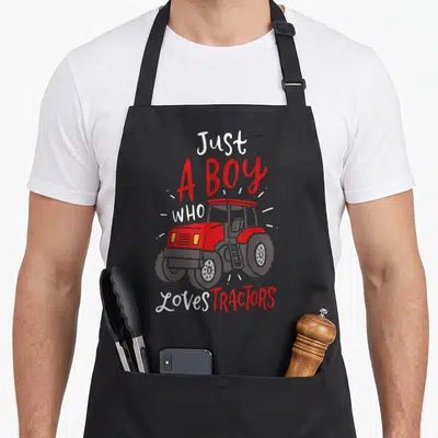 Funny tractor apron - Red and grey cartoon tractor with Just A Boy Who Loves Tractors text — shown on a black cotton canvas apron against a white background