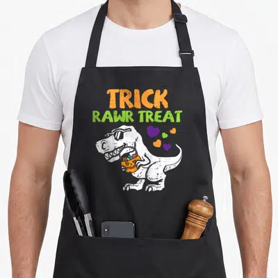 Rawr halloween apron - Cartoon dinosaur in sunglasses holding pumpkin bucket with hearts and trick rawr treat text — shown on a black cotton canvas apron against a white background