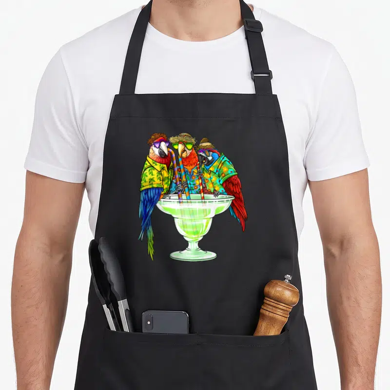 Parrot hawaiian apron - Three parrots in hats and sunglasses drinking from a large green cocktail glass with striped — shown on a black cotton canvas apron against a white background