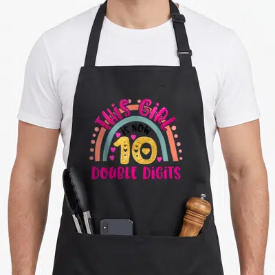 Funny double birthday apron - Rainbow graphic with pink text THIS GIRL IS NOW 10 DOUBLE DIGITS and gold number — shown on a black cotton canvas apron against a white background