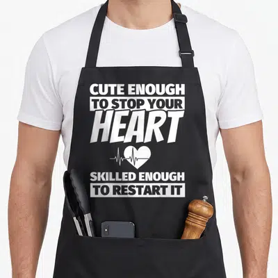 Nursing school nurse apron - White stacked text reading cute enough to stop your heart skilled enough to restart it — shown on a black cotton canvas apron against a white background