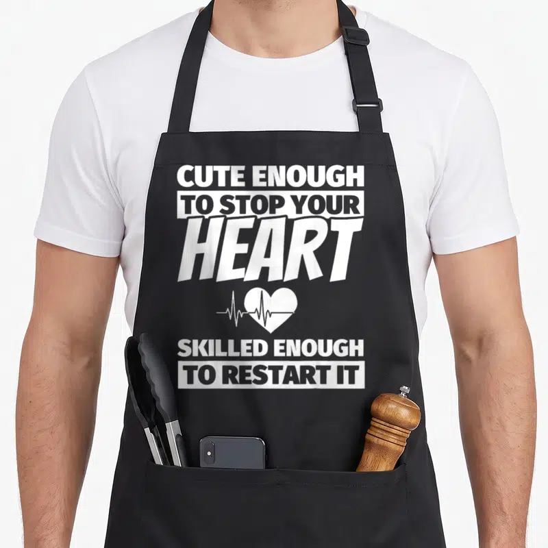 Nursing school nurse apron - White stacked text reading cute enough to stop your heart skilled enough to restart it — shown on a black cotton canvas apron against a white background