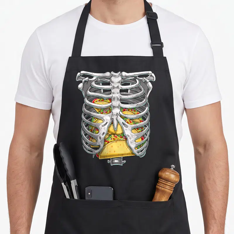 Skeleton rib halloween apron - Cartoon white ribcage filled with yellow, green, and red tacos in a cluster — shown on a black cotton canvas apron against a white background