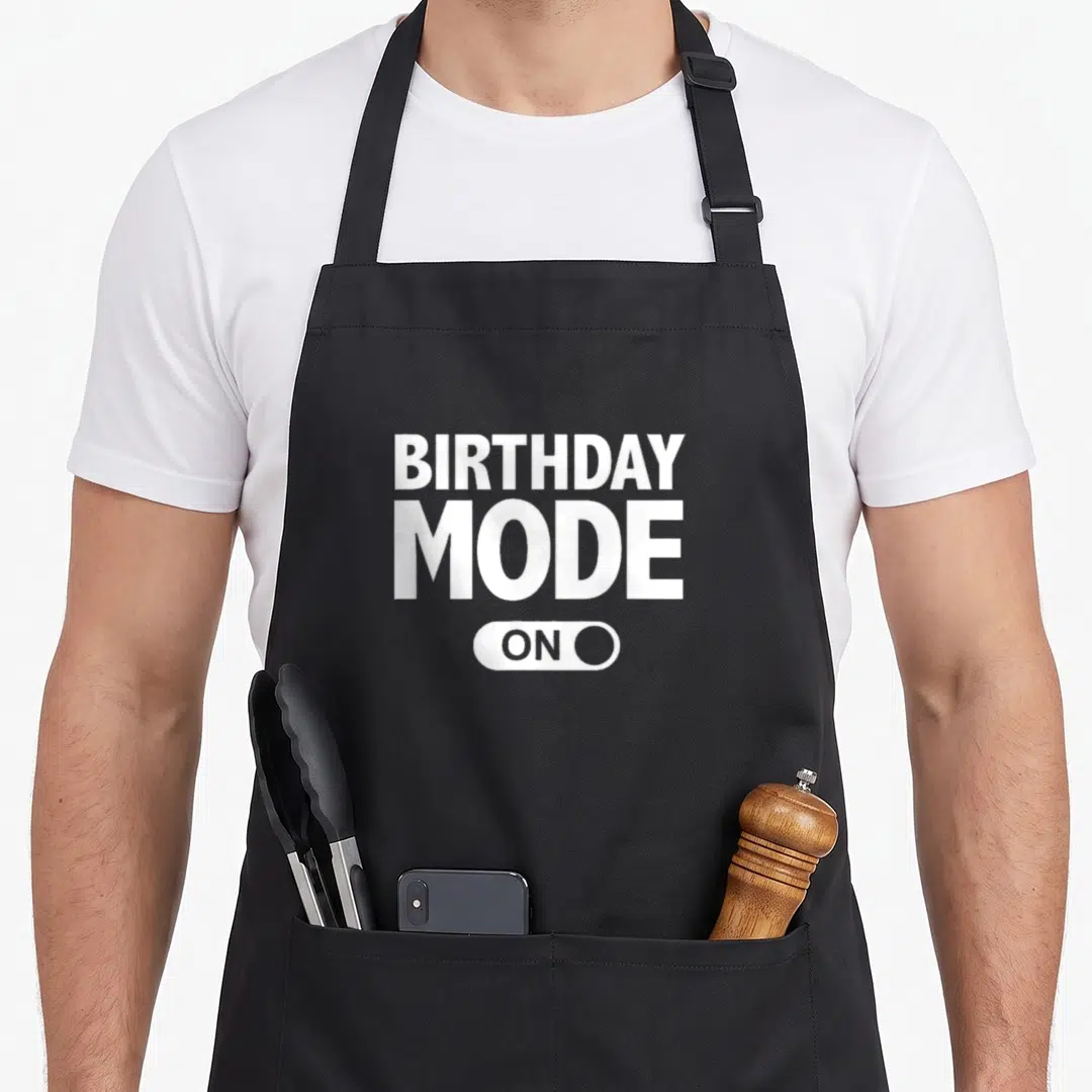 Mode birthday apron - BIRTHDAY MODE ON text inside a white toggle switch graphic on grey — shown on a black cotton canvas apron against a white background