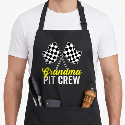 Pit grandma apron - Crossed black and white checkered flags above yellow Grandma and white PIT CREW text — shown on a black cotton canvas apron against a white background