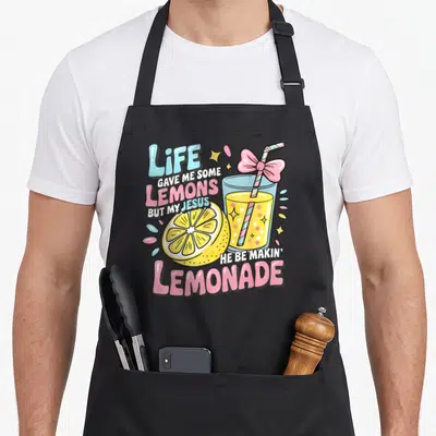 Makin lemonade apron - Cartoon lemonade glass with pink bow and life gave me lemons text — shown on a black cotton canvas apron against a white background