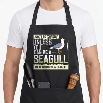 Seabird bird apron - Cartoon seagull overlapping stacked text Always Be Yourself Unless You Can Be a Seagull — shown on a black cotton canvas apron against a white background