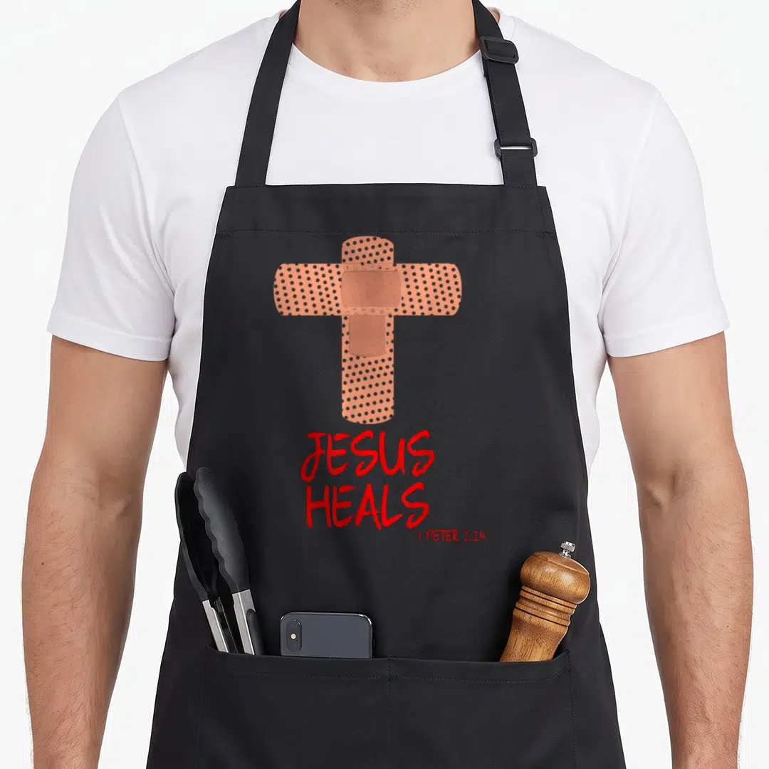 Heal nurse apron - Tan bandage cross above red JESUS HEALS 1 Peter 2:24 text — shown on a black cotton canvas apron against a white background