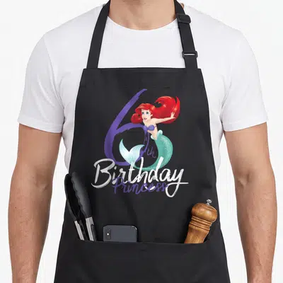 Pose princess apron - Mermaid overlapping purple 6 with 6th Birthday Princess text in white and purple script — shown on a black cotton canvas apron against a white background