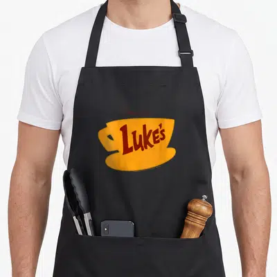 Luke gilmore girls apron - Yellow coffee cup with red Luke's text and Gilmore girls label above — shown on a black cotton canvas apron against a white background