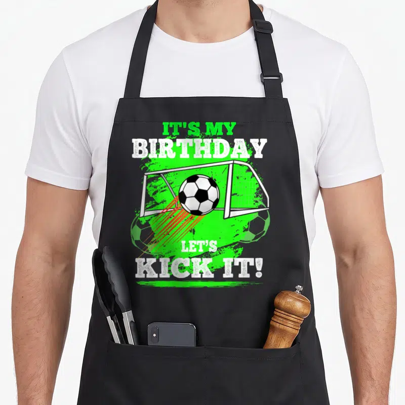 Kick birthday apron - Soccer ball hitting goal net with green splatter and IT'S MY BIRTHDAY LET'S KICK IT! text — shown on a black cotton canvas apron against a white background