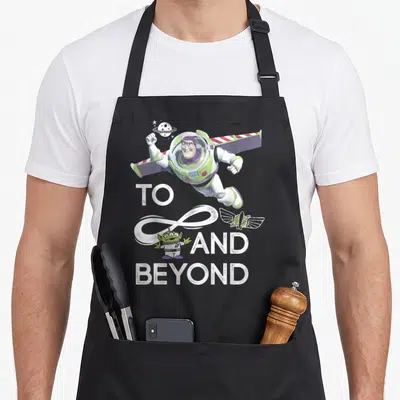 Alien infinity apron - Flying Buzz Lightyear and green alien surrounding white TO AND BEYOND text with infinity symbol — shown on a black cotton canvas apron against a white background