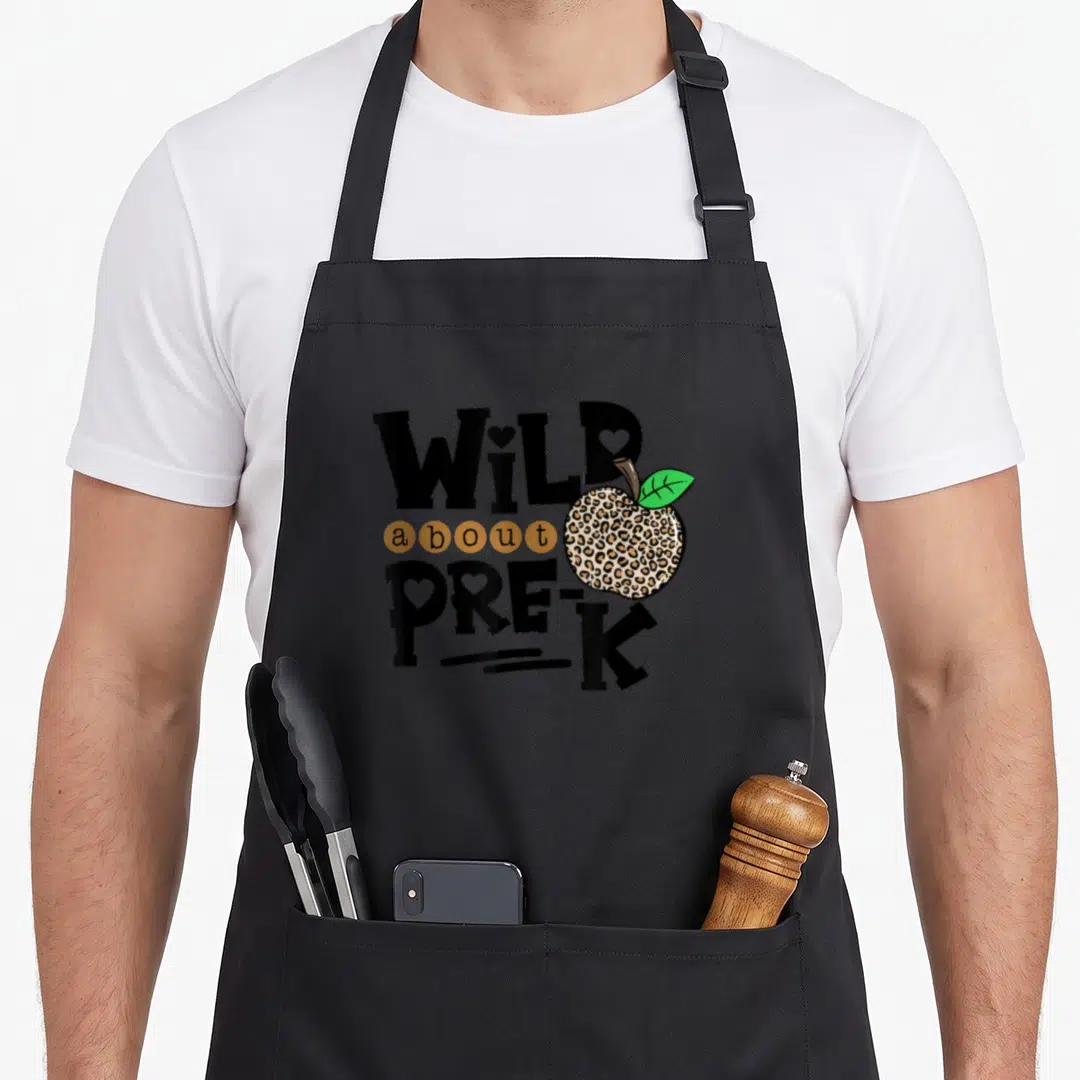 Wild pre teacher apron - Leopard print apple overlapping WILD about PRE-K text with hearts in letters — shown on a black cotton canvas apron against a white background