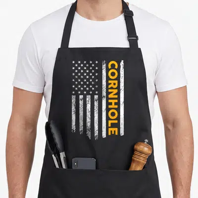 Cornhole us aprons - Vertical distressed white USA flag with orange CORNHOLE text overlaid on stripes — shown on a black cotton canvas apron against a white background