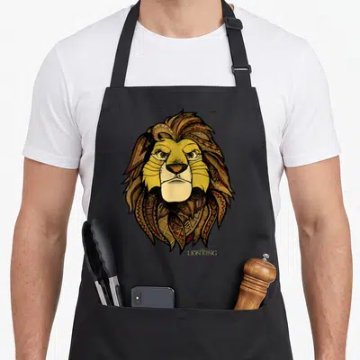 Tribal lion apron - Line art lion head with tribal mane patterns and THE LION KING text — shown on a black cotton canvas apron against a white background