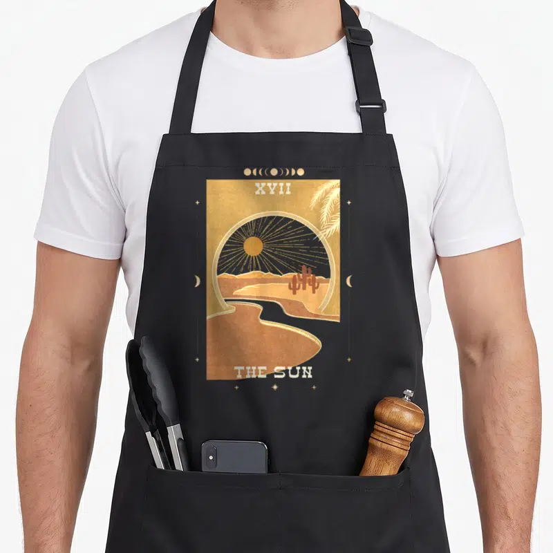 Dark boho apron - XVII THE SUN tarot desert scene with sun cactus river moon phases stars in tan brown yellow white — shown on a black cotton canvas apron against a white background