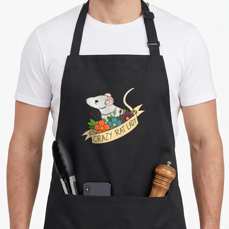 Flower rodent pet apron - Cartoon white rat behind orange blue and purple flowers on beige ribbon — shown on a black cotton canvas apron against a white background