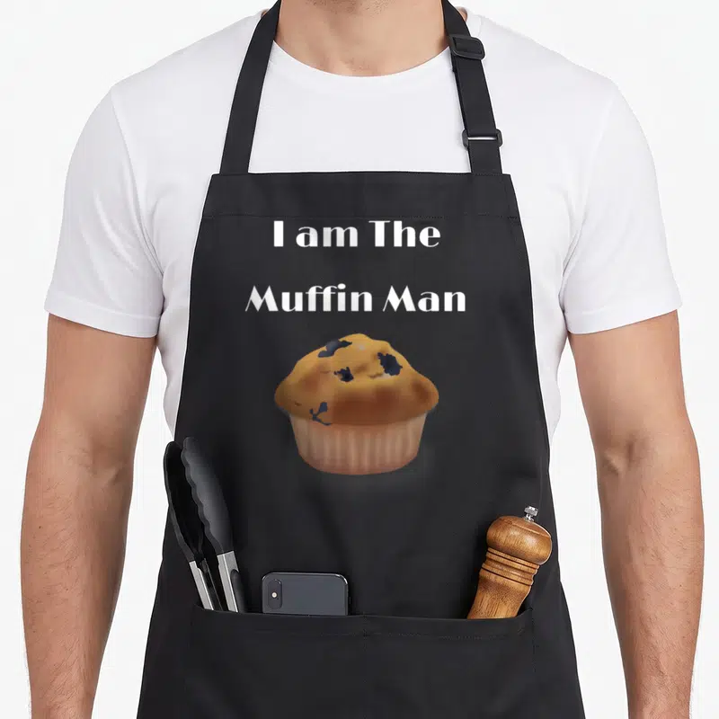 Funny muffin man apron - I am The Muffin Man text above a cartoon blueberry muffin in brown and blue — shown on a black cotton canvas apron against a white background