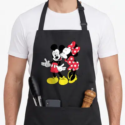 Funny mouse lover apron - Mickey and Minnie standing side-by-side cartoon kiss in black red yellow white — shown on a black cotton canvas apron against a white background